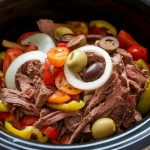 a close-up of a slow cooker (crockpot) filled with shredded beef, sliced bell peppers, cherry tomatoes, onions, and olives.