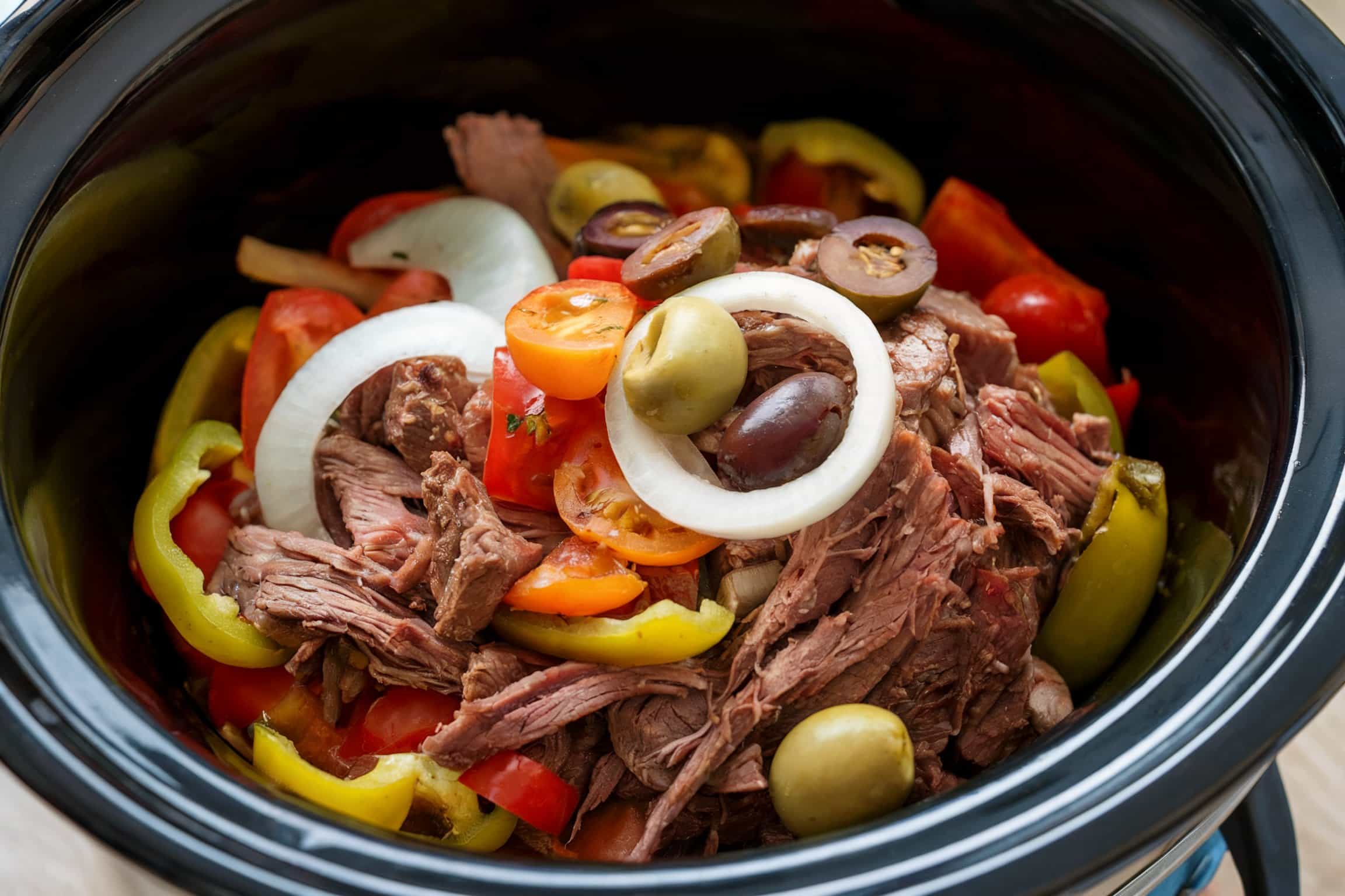 a close-up of a slow cooker (crockpot) filled with shredded beef, sliced bell peppers, cherry tomatoes, onions, and olives.