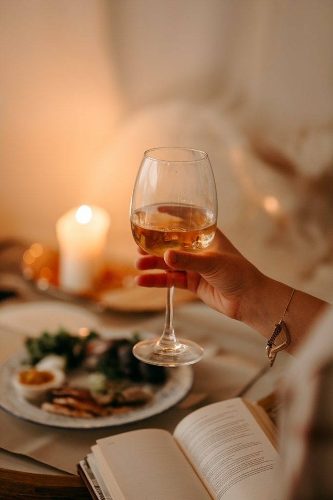 Person holding a glass of white wine with a plate of food and a book in the background