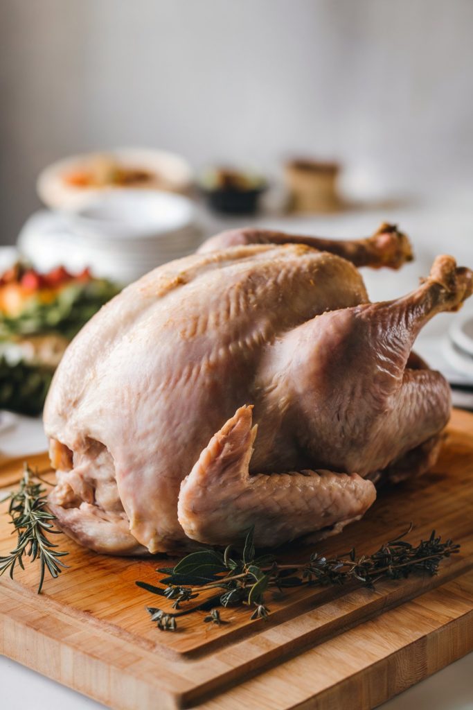 A whole raw turkey is placed on a wooden cutting board, surrounded by fresh onions, garlic, parsley, black peppercorns, and green herbs