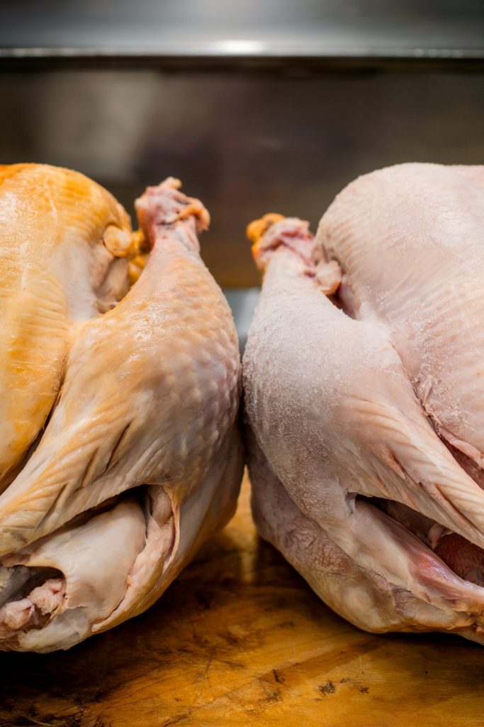Two whole raw turkeys are placed side by side on a wooden surface, one fresh and the other frozen with visible frost on its surface