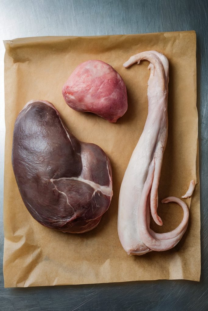 Three raw organ meats, including a dark liver, a pinkish gizzard, and a long, white turkey neck, are arranged on a sheet of parchment paper on a metal countertop