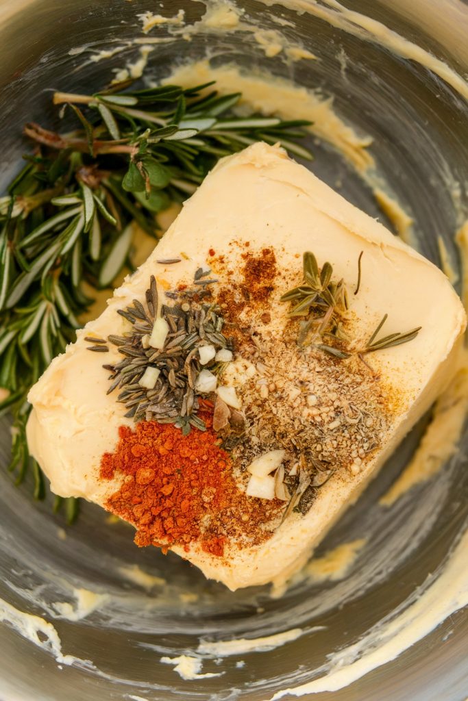 A close-up of a slab of softened butter in a mixing bowl, topped with fresh rosemary and an assortment of spices, including paprika, dried herbs, and minced garlic