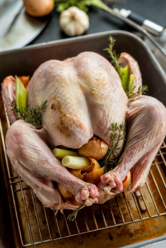 A raw turkey sitting on a wire rack in a roasting pan, stuffed with onions, celery, and fresh rosemary sprigs