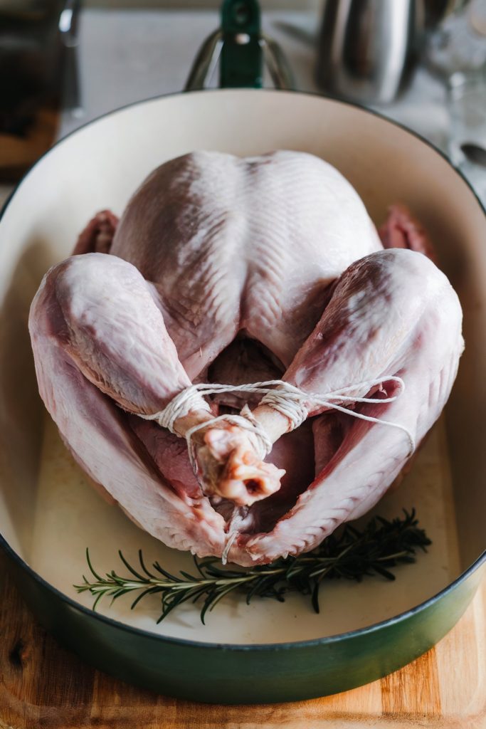 A raw, whole turkey with its legs tied together with twine, placed in a large green roasting pan with sprigs of fresh rosemary underneath