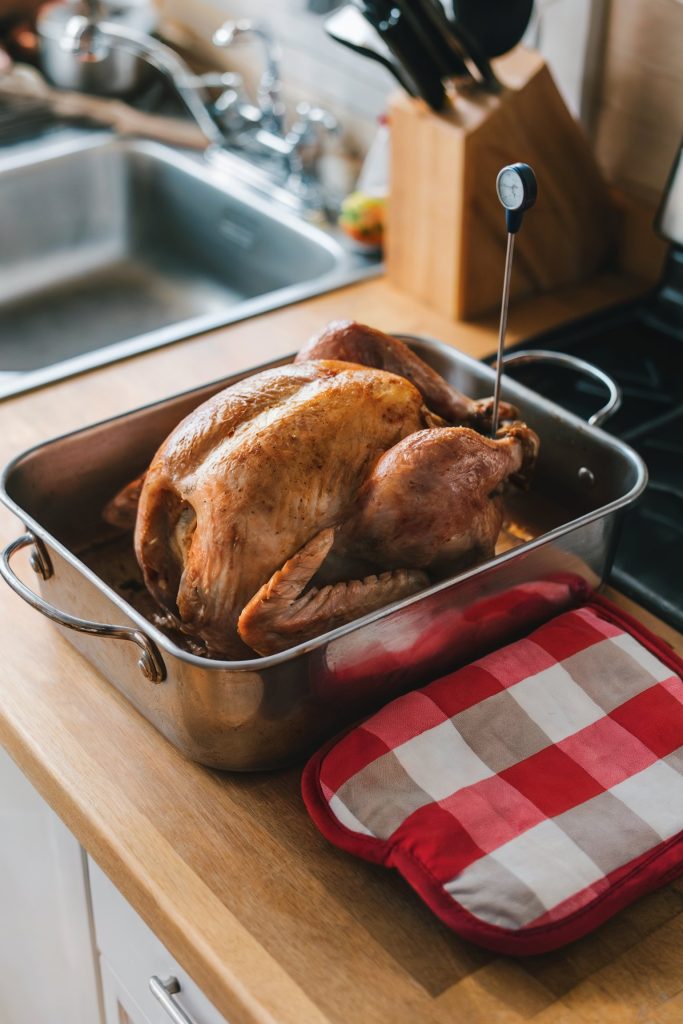 A freshly roasted turkey resting in a roasting pan on a kitchen countertop. A meat thermometer is inserted into the turkey, and a red-and-white checkered oven mitt lies nearby