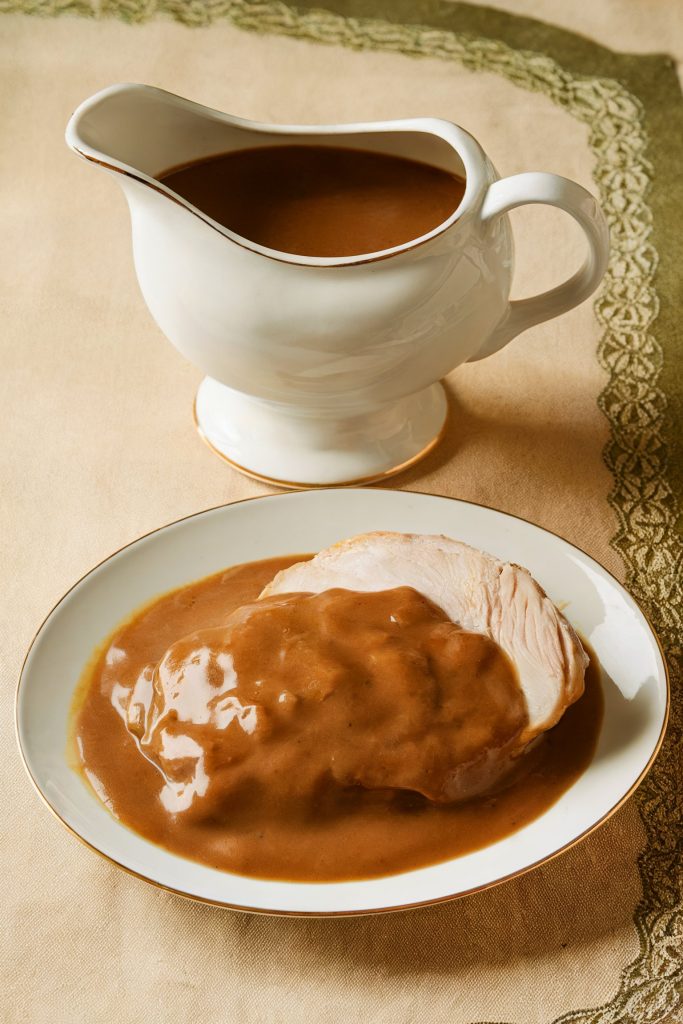A slice of turkey with gravy on a plate and a gravy container beside it