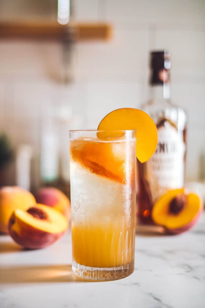 Indoor countertop view of a highball glass with golden peach cocktail, peach slice on rim, no text or logos.