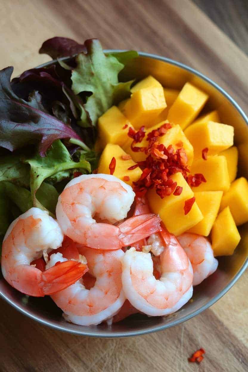 Indoor shot of a shallow bowl holding cooked pink shrimp, diced mango, mixed greens, and a sprinkling of red chili flakes. No text or logos present