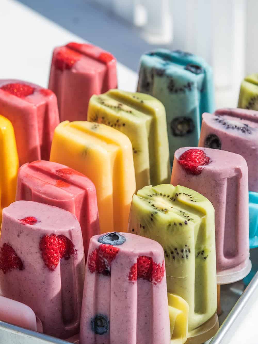 Three colorful smoothie popsicles on a wooden surface with fresh fruits around them.