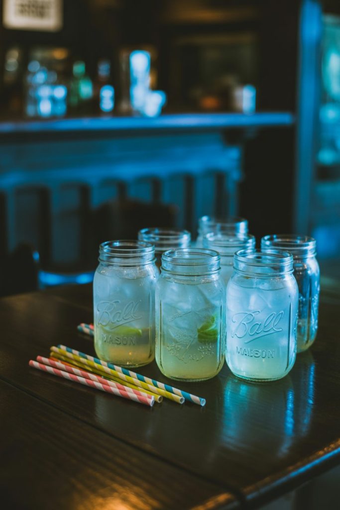 Indoor dimly lit scene with mason jars of tonic-water lemonade under black light, emitting a ghostly blue glow; paper straws, no logos.