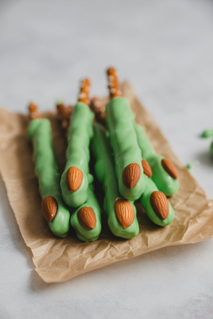 Photo of green candy-coated pretzel rods with almond “nails,” indoors on parchment. No text or logos.