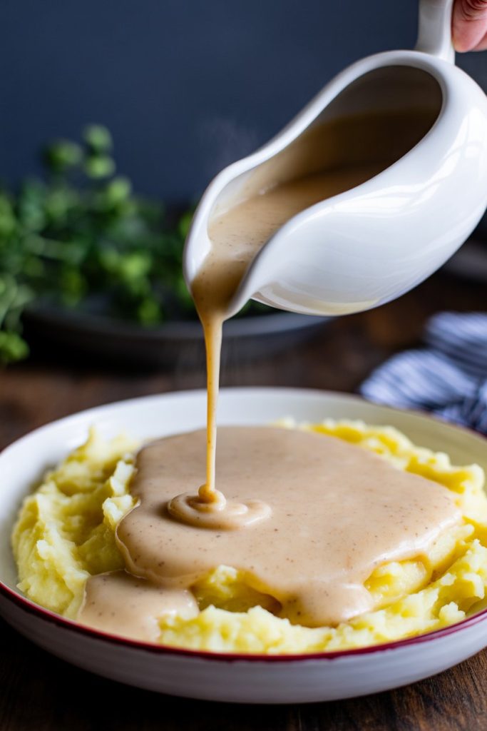 A gravy boat pouring creamy off-white cauliflower gravy over mashed potatoes, indoors. No text or logos.