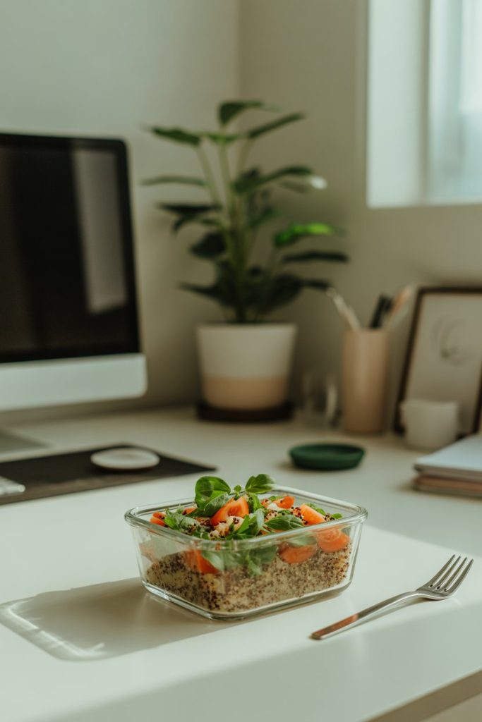 An indoor office desk with a glass container of quinoa salad, small fork on the side—photo, no branding or logos.