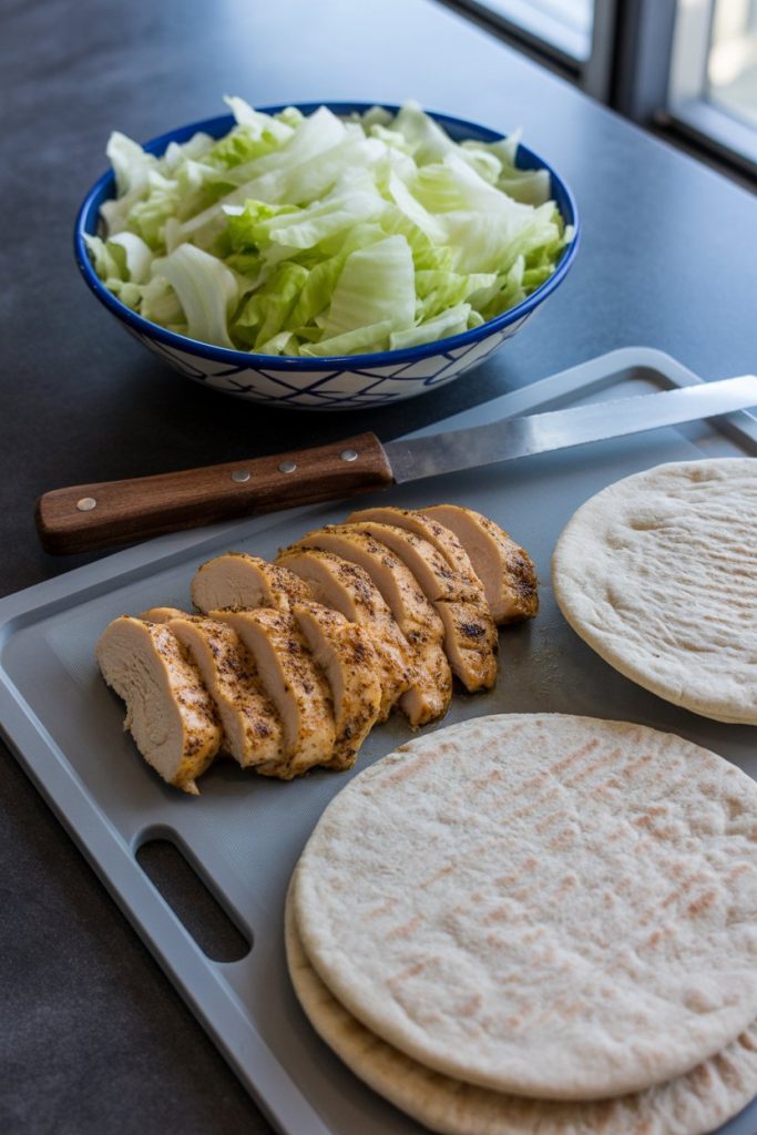 An indoor prep board holding sliced grilled chicken, whole-wheat pita halves, and shredded lettuce ready for assembly. No text or logos present.