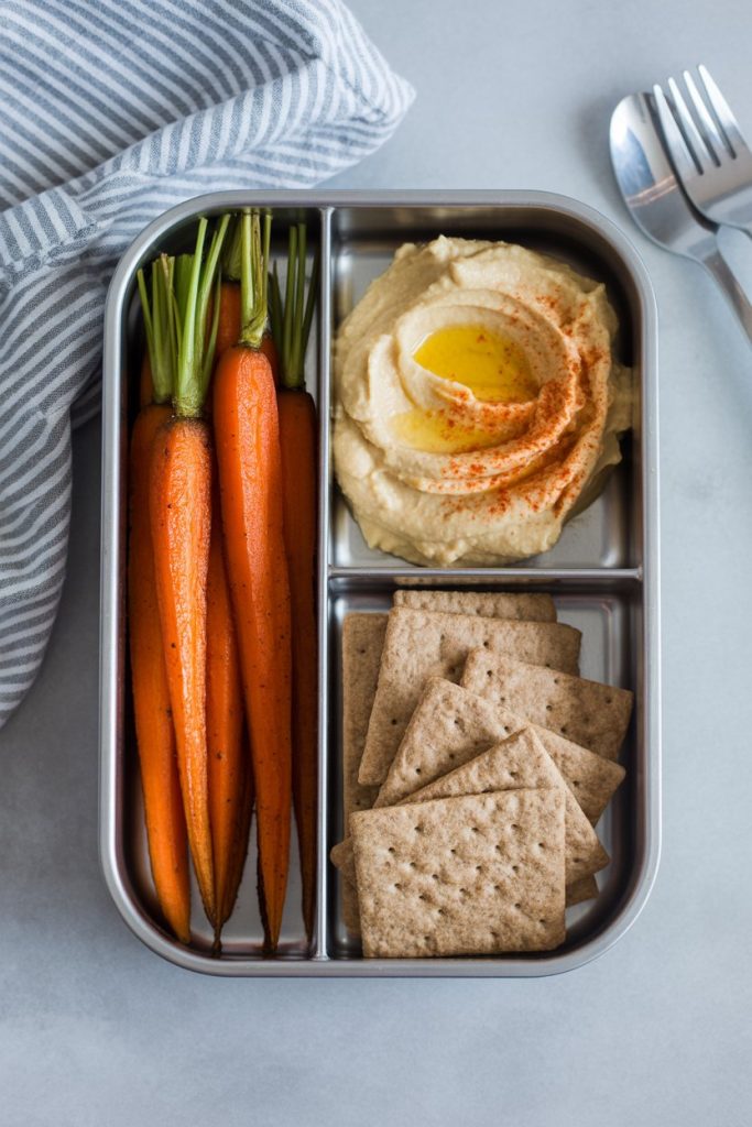 Indoor meal prep container holding harissa-spiced roasted carrots, a scoop of classic hummus, and whole-grain crackers. No text or logos present.