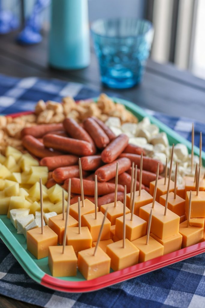 Indoor photo of a colorful plastic platter featuring cheddar cubes with toothpicks, cocktail sausages, pineapple chunks, and cheese balls; no logos or text.