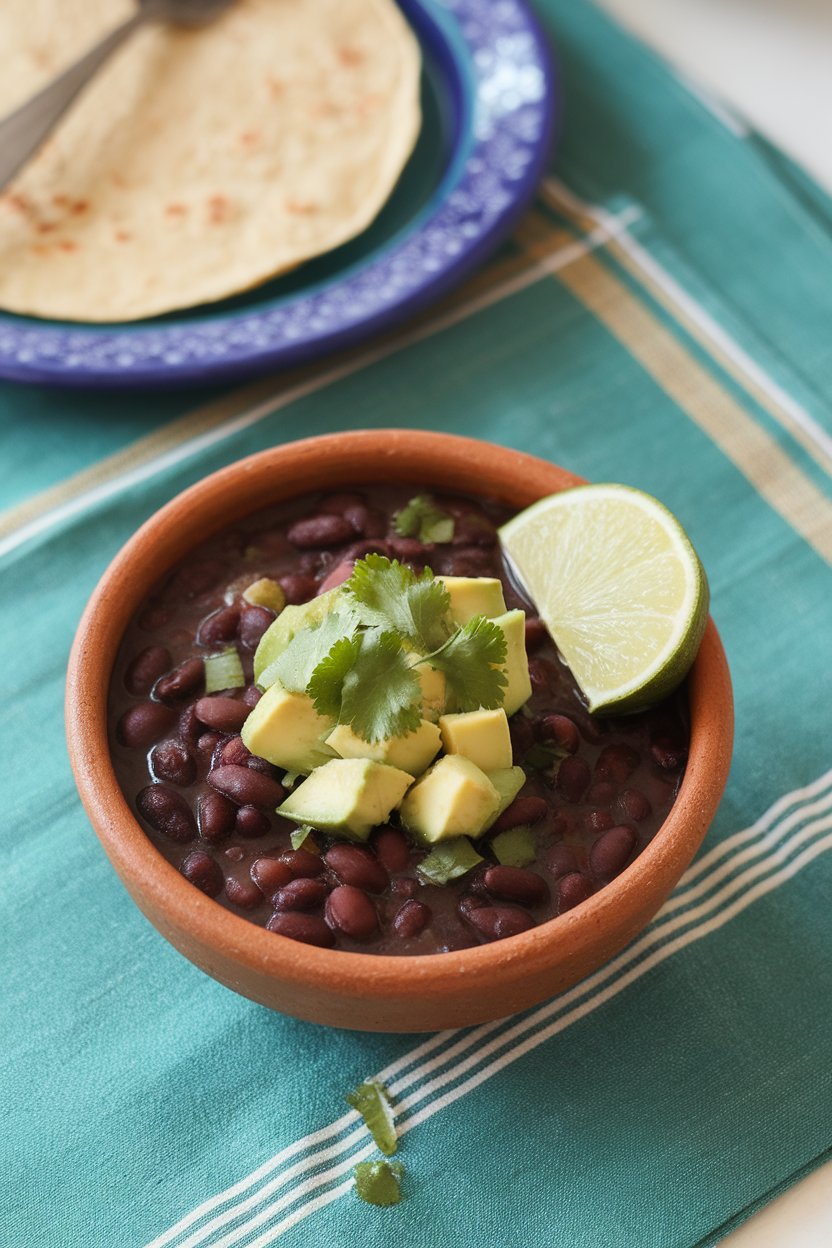 Indoor tabletop with a clay bowl of black bean soup, topped with diced avocado, cilantro, and a lime wedge. No text or logos. Photo.