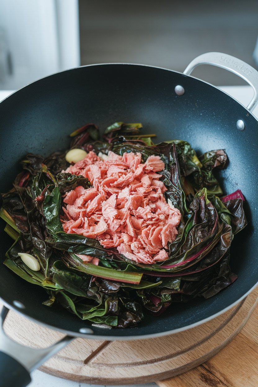 Wok of sautéed Swiss chard ribbons and tuna flakes with garlic cloves visible; indoor kitchen light, no branding.
