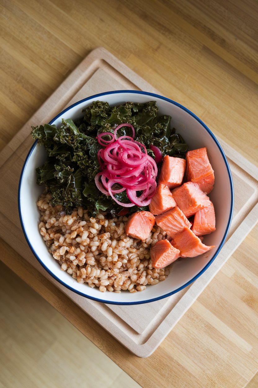 An indoor countertop bowl containing cooked farro, massaged kale, roasted salmon chunks, and pickled red onions, photographed overhead. No text or logos anywhere.