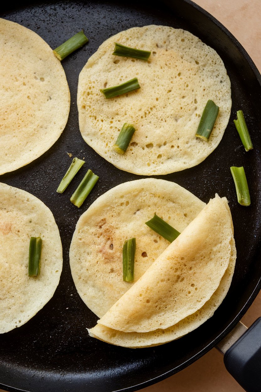 An indoor griddle shot of thick adai pancakes speckled with green drumstick leaves, one folded over. No text or logos. Photo, not illustration.