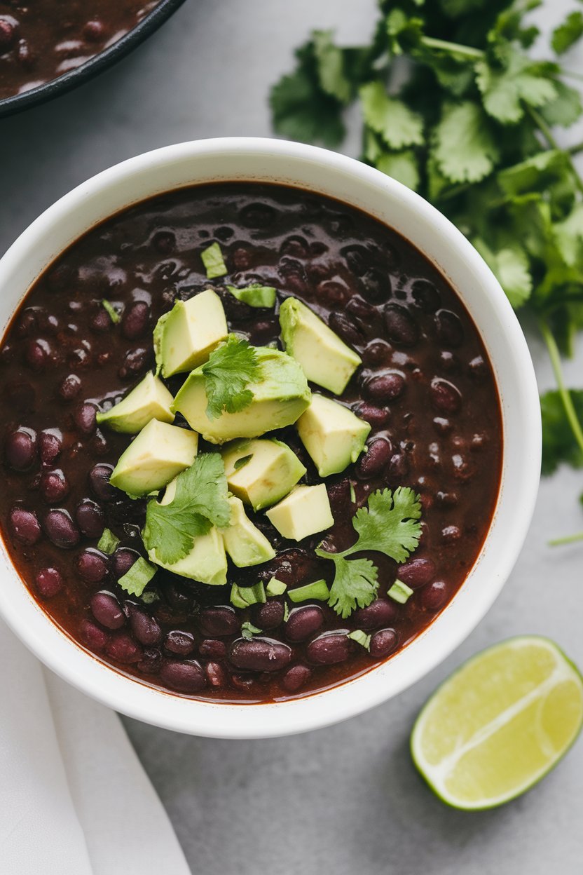 Indoor photo of dark black bean soup topped with diced avocado and cilantro, lime wedge on the side; no text or logos