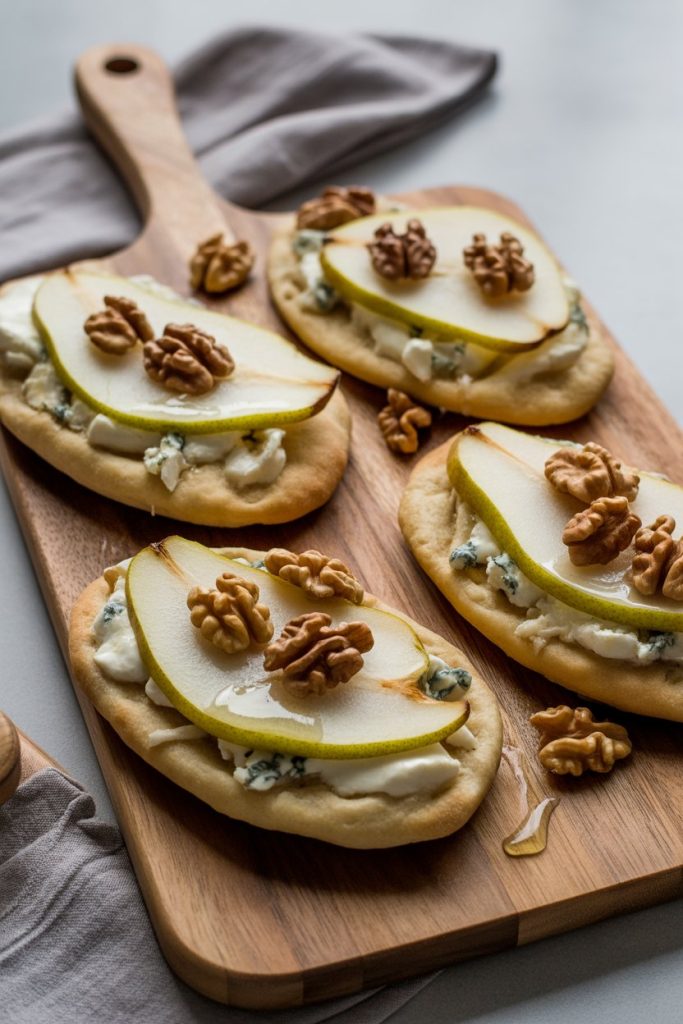 A wooden indoor serving board holding oval flatbreads topped with thin pear slices, melted Gorgonzola, and toasted walnuts, drizzled with honey. No text or logos visible.