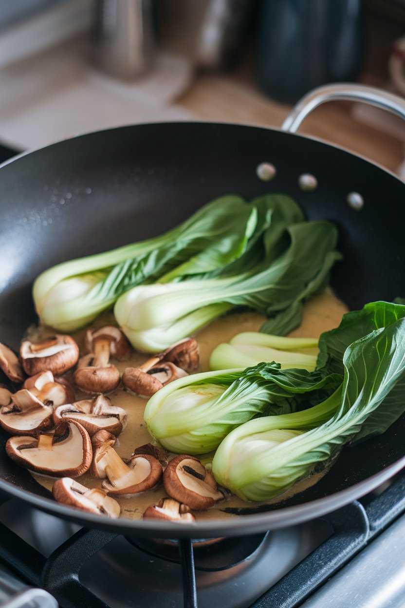 Indoor photo of a wok containing bright bok choy leaves and browned mushrooms in light sauce; soft stovetop light, no text or logos
