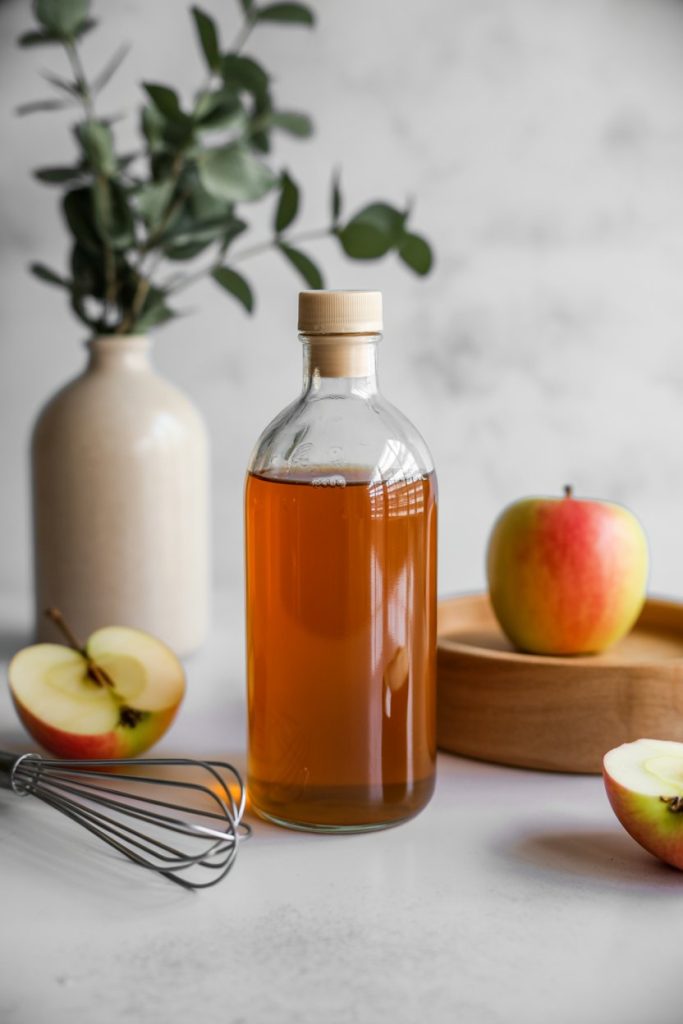 Indoor clear glass filled with cloudy apple cider vinegar beside a small whisk; no text or logos. Photo.