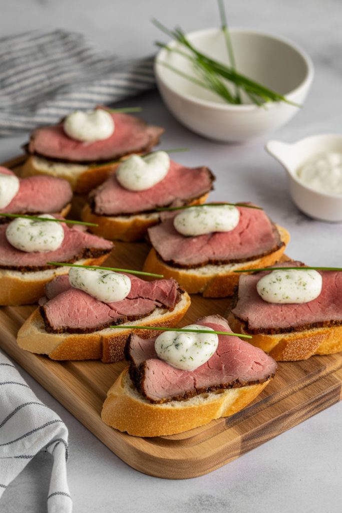 Photo of an indoor serving board topped with toasted baguette slices, each holding rare roast beef, a dollop of horseradish cream, and chives. No text or logos anywhere.