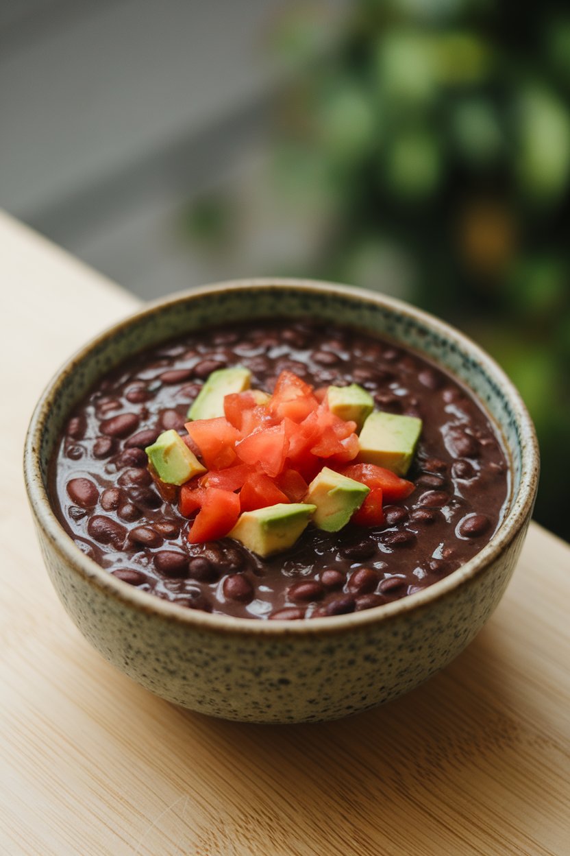 Indoor bowl filled with thick black bean soup topped with diced tomato and avocado cubes; no text or logos; photo.