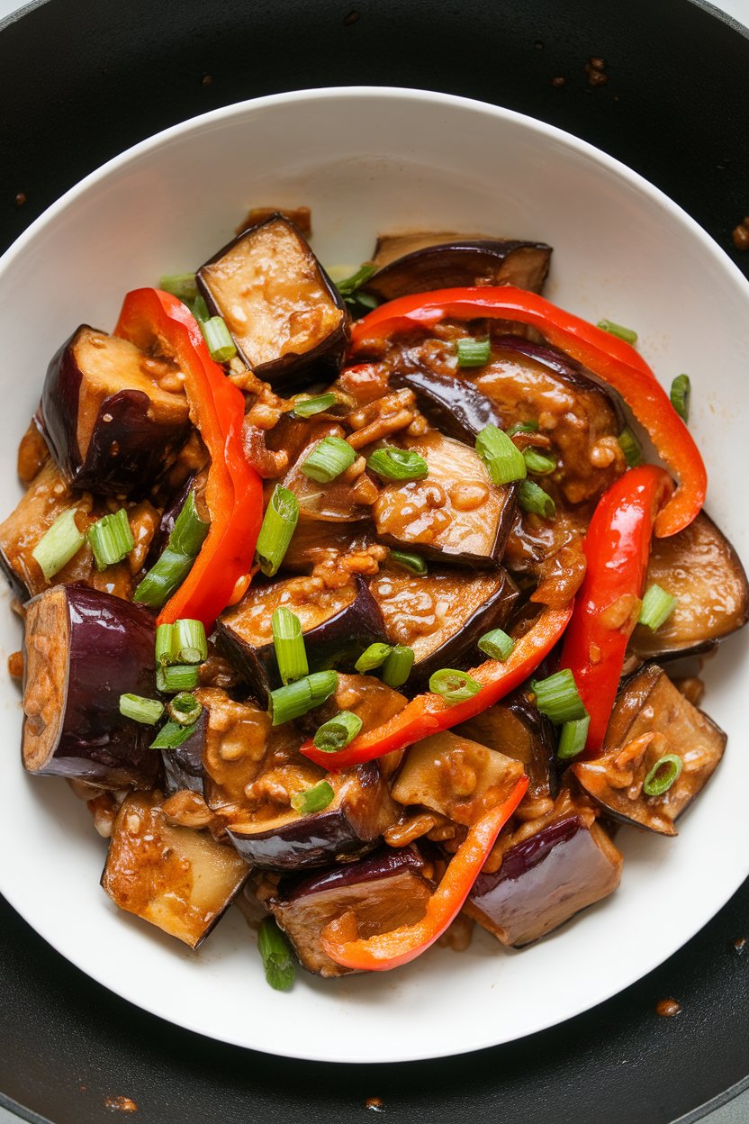 Indoor wok shot of tender eggplant cubes coated in a glossy garlic sauce with bell pepper strips and scallions. No text or logos. Photo only.